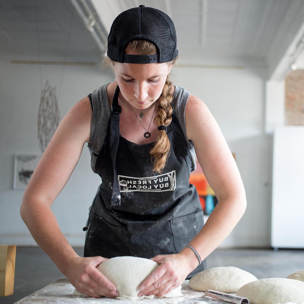 Photo of a young white woman with a black hat, worn backwards, and a long braid is kneading dough on a table